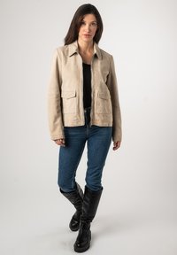 Beige Wildlederjacke mit einem Kragen, zwei Fronttaschen und einem Reißverschluss. Über einem schwarzen Oberteil getragen und kombiniert mit blauen Jeans und schwarzen Stiefeln.