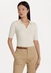 Ριχτό κρεμ πόλο μπλουζάκι με V-ντεκολτέ και κοντά μανίκια, συνδυασμένο με μπεζ παντελόνι και καφέ ζώνη. Λείος υφασμάτινος χαρακτήρας και εφαρμοστό σχέδιο.