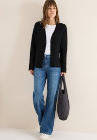 Cardigan nero lavorato a maglia con scollatura a V e bottoni, abbinato a jeans a gamba larga blu. Il modello tiene una borsa a spalla grigia, indossando sneakers bianche.