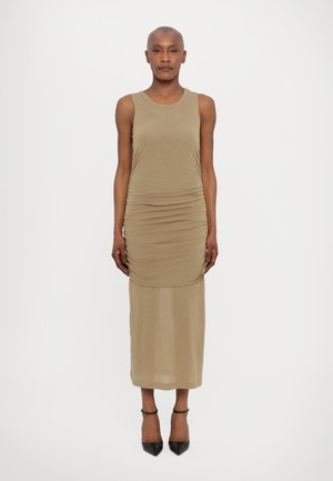 Robe fitted kaki avec un design sans manches, tissu texturé, détails froncés sur les côtés et une couche inférieure semi-transparente lisse. Escarpins noirs à talons aiguilles.