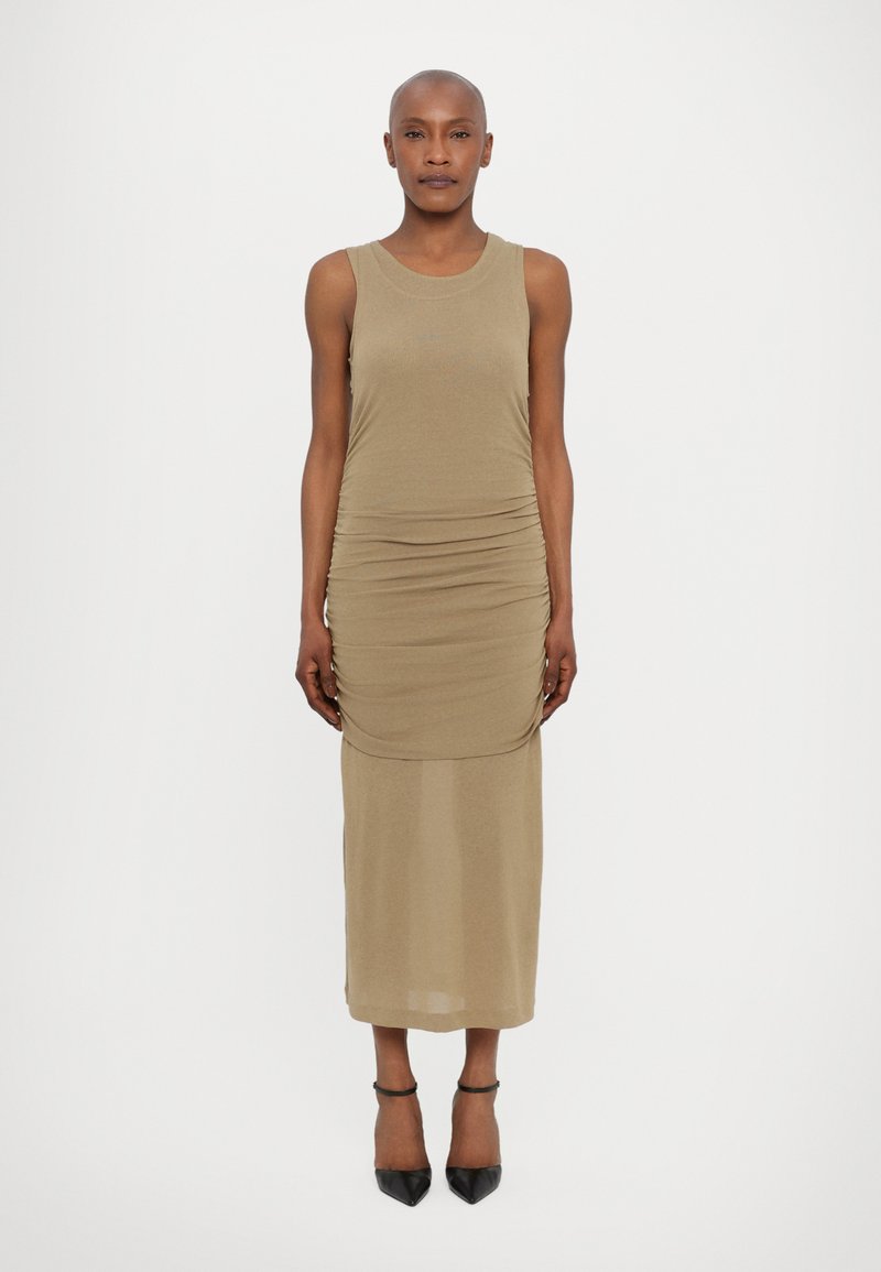 Rochie kaki ajustată cu un design fără mâneci, fabrică texturată, detalii strânse pe lateral și un strat inferior semi-transparent, neted. Tocuri stiletto negre.