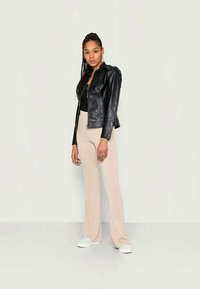 Jaqueta de couro sintético preta com fecho de zip, combinada com calças largas de malha bege claro e ténis brancos. Modelo em pé contra um fundo liso.