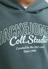 Felpa verde con la scritta "JACK & JONES Coll. Studio Creata dal team J&J dal 1990" stampata in bianco sul petto.