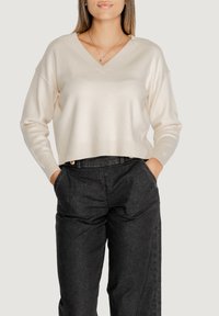 Maglione beige chiaro a V con maniche lunghe, realizzato in materiale morbido, di lunghezza cropped, abbinato a pantaloni grigi scuri a vita alta.