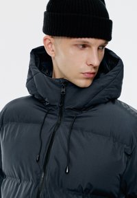 Czarna kurtka puchowa z wysokim kołnierzem, dwoma przednimi kieszeniami i zapięciem na zamek z przodu. Tkanina wydaje się gładka z lekko błyszczącym wykończeniem.