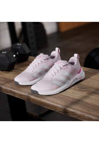 Svetlo rožnate športne superge iz mrežastega materiala s tremi belimi črtami, teksturiranim prstom in sivo podplatom, prikazane na leseni površini.