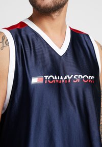 Canotta sportiva della Marina con rifiniture bianche e accenti rossi. Presenta il logo "TOMMY SPORT" in grassetto rosso e bianco sulla parte centrale.