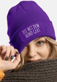 Purple knit beanie with a rectangular patch displaying the text "DIE MIT DEM HUND GEHT" in white. Ribbed texture and snug fit.