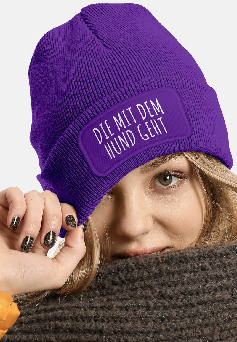 Purple knit beanie with a rectangular patch displaying the text "DIE MIT DEM HUND GEHT" in white. Ribbed texture and snug fit.