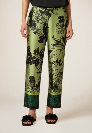 Grüne Satin-Hose mit schwarzem tropischem Blumenmuster und dunkelgrünen Bündchen, getragen mit schwarzen Sandalen mit Blumenmuster auf hellem Hintergrund.