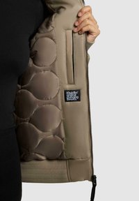 Giacca imbottita beige con orlo a coste, motivo trapuntato testurizzato all'interno e una piccola etichetta in tessuto con il nome del brand.