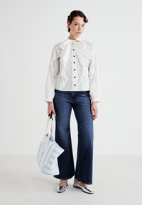 Blusa blanca con acentos de volantes y botones negros, combinada con jeans de mezclilla oscuros de campana. Zapatos planos plateados y un bolso de algodón a rayas azul claro.