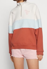 Färgblockerad sweatshirt med hög krage, har sektioner i vitt, ljusblått och rostorange. Broderad logotyp i rosa. Mjuk tyg.