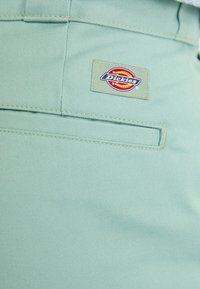 Pantalones verde claro hechos de una tela duradera que presentan un bolsillo trasero con un parche rectangular del logo de Dickies. Diseño simple con detalles de costura.