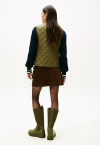 Gepatchte olivgrüne Weste über einem marineblauen Strickpullover, kombiniert mit einem braunen Cordminirock und grünen Gummistiefeln bis zu den Knien.