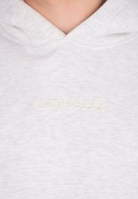 Gros plan sur un hoodie gris clair avec un texte brodé subtil "DROPSIZE" sur la poitrine et la peau du cou et du haut de la poitrine visibles.