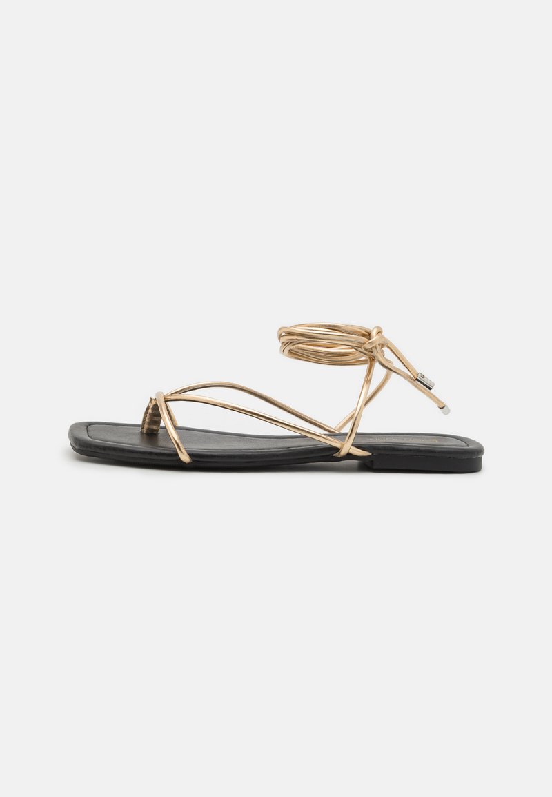 Black flat sandal featuring thin gold straps that wrap around the ankle and a minimalist design with a smooth, rectangular footbed.