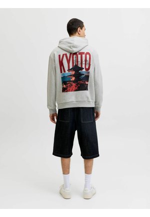 Homme portant un sweat à capuche gris avec un grand texte "KYOTO" et un graphique de pagode au dos, un short noir, des chaussettes blanches et des baskets blanches, debout de dos.