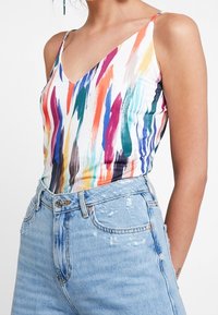 Kolorowy top bez rękawów z wzorami przypominającymi wielokolorowe pociągnięcia pędzla, wykonany z lekkiego materiału, zestawiony z podniszczonymi jasnoniebieskimi dżinsami z wysokim stanem.