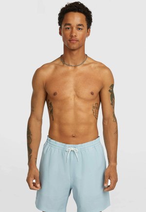 Homme torse nu avec des cheveux bouclés portant un short de bain bleu clair, un collier de perles et des boucles d'oreilles, montrant plusieurs tatouages sur les bras et le torse.