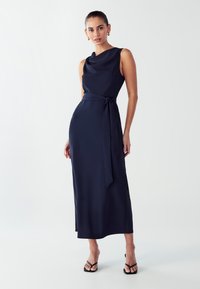 Tamno plava haljina bez rukava s šal ovratnikom, pojasom u struku i ravnim midi siluetom. Glatka tekstura s sjajnim završetkom.