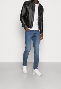 Sort læderjakke med lynlåslukning, hvid t-shirt, blå jeans og hvide sneakers, båret af en person mod en ensfarvet baggrund.