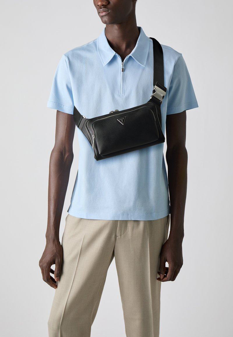 Borsa a tracolla in pelle nera con chiusura a zip e dettaglio del logo, indossata a tracolla sopra una polo azzurro chiaro. Tracolla regolabile.