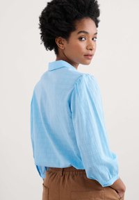 Svetlo modra, teksturirana bluza s ovratnikom in nabranimi rokavi, v kompletu s smešnimi hlačami rjave barve z elastičnim pasom. Zadnji pogled prikazuje dolžino in slog majice.