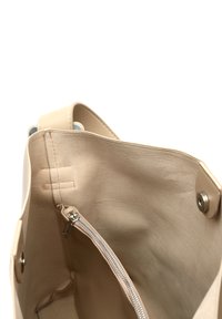 Beige kantokassi, jolla on sileä pinta, avoin yläosa ja sisäinen vetoketjullinen tasku. Varustettu metallisilla yksityiskohdilla ja minimalistisella muotoilulla.