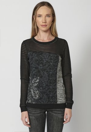 Pull en maille noire avec un design en maille texturé, motif floral sur un panneau, et tissu gris contrastant. Col rond et poignets côtelés.