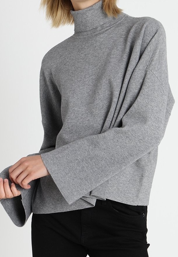 Personne portant un pull à col roulé gris ample avec des manches larges et un pantalon noir, ajustant le poignet d'une manche.