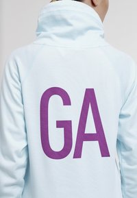 Sudadera azul clara con cuello alto, que presenta un gran "GA" morado impreso en la espalda. Tejido suave con una textura lisa.