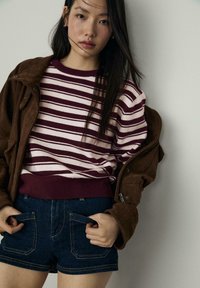 Sweter w paski w kolorze burgundowym i kremowym, połączony z ciemnoniebieskimi szortami dżinsowymi. Brązowa zamszowa kurtka nałożona na sweter. Dwie widoczne kieszenie z przodu.