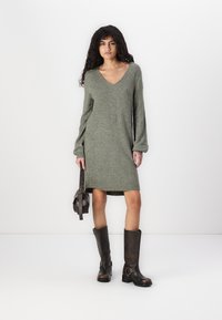 Robe en tricot verte olive, longueur genou, avec un décolleté en V et des manches longues. Associée à des bottes en cuir marron hautes avec des boucles.