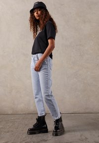 Jeune femme aux cheveux bouclés portant une casquette noire, un t-shirt noir, un jean bleu clair et des bottes noires à plateforme, debout contre un mur uni.