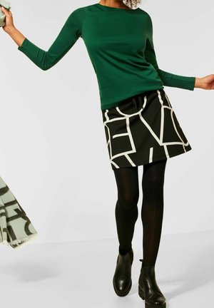 Femme portant un haut vert à manches longues, une jupe noire avec un motif abstrait blanc, des collants noirs et des bottines noires, debout les bras écartés.