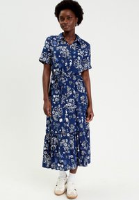 Mujer de pie con un vestido midi azul con estampado floral, abotonado, de manga corta y bajo con volantes, llevando calcetines blancos y zapatillas deportivas beige.