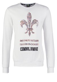 Bílá mikina s dlouhým rukávem, na které je barevný květinový motiv fleur-de-lis a černý textový prvek. Měkká látka s žebrovanými manžetami.