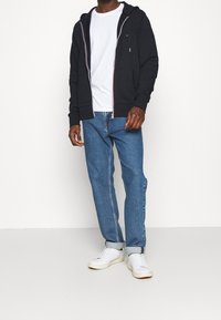 Sudadera negra con cremallera y capucha con cordón, camiseta blanca debajo, jeans azules y zapatillas blancas. Ajuste casual, puños acanalados y cinturilla.