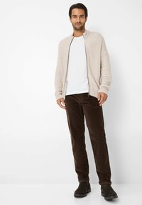Beige gerippter Cardigan, weißes T-Shirt, braune Cordhosen und schwarze Stiefeletten. Schlichtes Design, Reißverschluss vorne und hochgekrempelte Hosen.