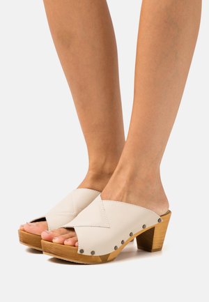 Mules ouvertes en cuir blanc avec un design entrecroisé, talon en bloc de bois et clous en métal sur le côté. Texture lisse et style minimaliste.