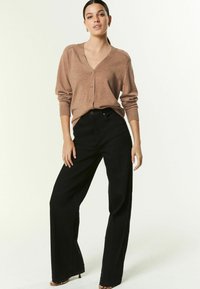 Cardigan en laine marron tricoté avec un décolleté en V et une fermeture à boutons, associé à un pantalon noir taille haute et large, mettant en avant une coupe décontractée.
