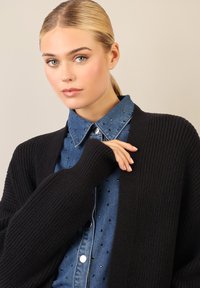 Cardigan nero a coste sopra una camicia di denim blu con dettagli in borchie argentate. Il cardigan presenta maniche ampie; la camicia ha la chiusura a bottoni e colletto.
