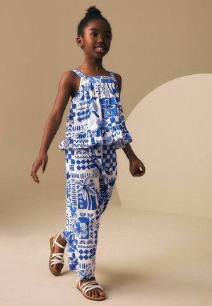 Young girl walking in blue and white patterned sleeveless top and matching pants with white braided sandals on neutral background.