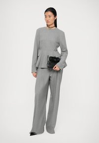 Femeie purtând un costum gri cu dungi fine, pantaloni cu volănaș, ținând o poșetă neagră texturată și o colier auriu în formă de inimă.