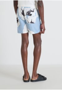 Costumi da bagno azzurri chiari con ampi motivi floreali bianchi e neri, con cintura elastica e una sola tasca sul retro. Indossati con sandali neri.