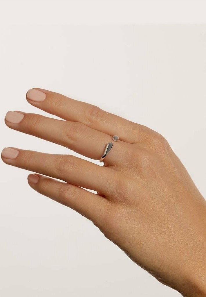 Main à la peau claire portant une bague argentée ouverte moderne avec deux extrémités arrondies, ongles peints d'une couleur nude douce.