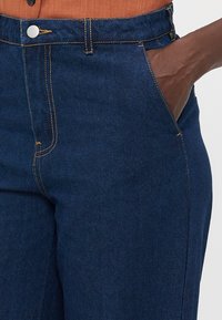 Mørkeblå denimbukser med oransje søm, med knappelukking, sidelommer og en glatt tekstur.