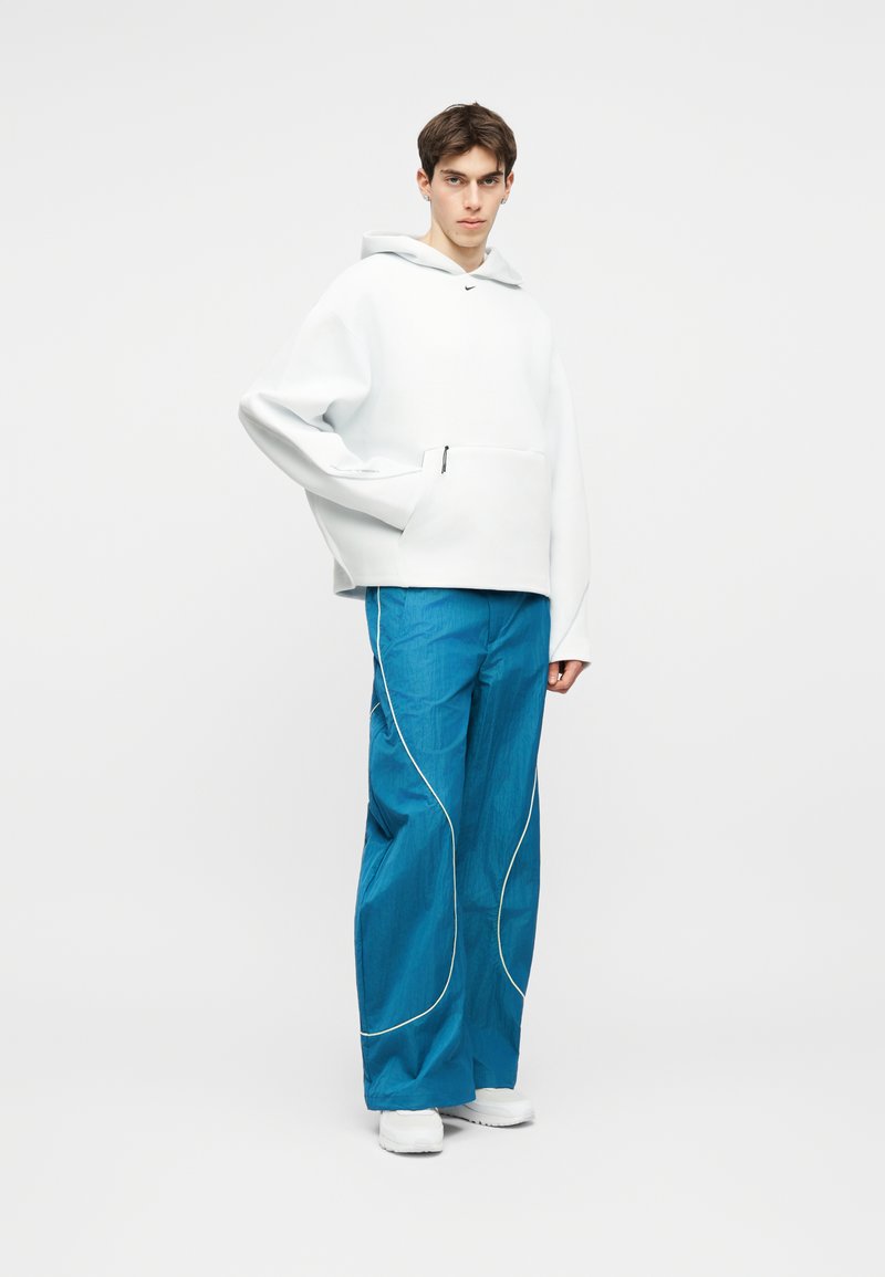 Giovane uomo in piedi che indossa una felpa oversize bianca e pantaloni larghi blu con piping bianco, insieme a scarpe da ginnastica bianche, su uno sfondo neutro.