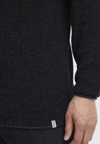 Schwarzer Strickpullover mit strukturiertem Muster, Rundhalsausschnitt und umgeschlagenen Säumen. Kleinere Markenlabel am Saum sichtbar.
